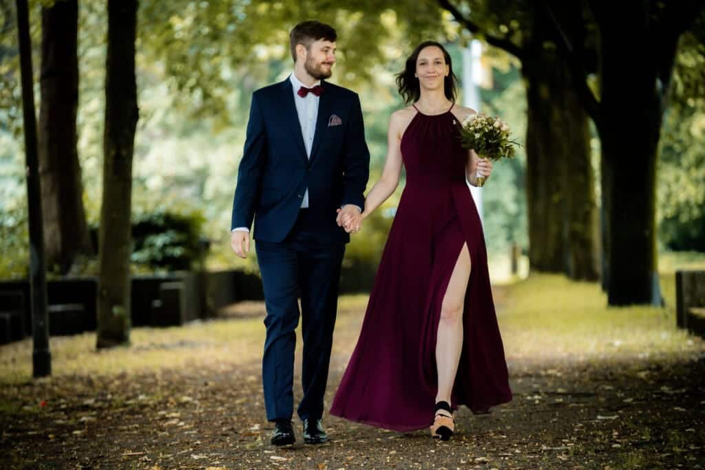 Ein Paar geht Hand in Hand über einen von Bäumen gesäumten Weg; die Frau trägt ein bordeauxrotes Kleid und einen Blumenstrauß