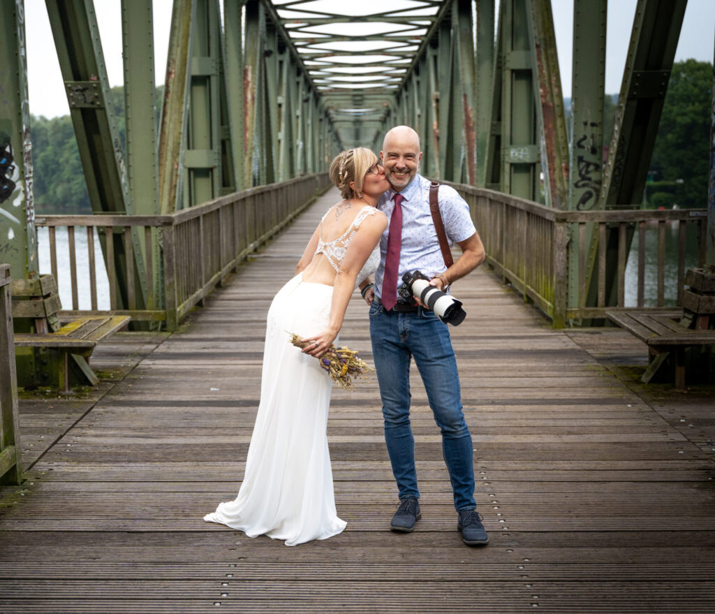 Braut küsst den Hochzeitsfotografen mit Kamera auf einer Holzbrücke
